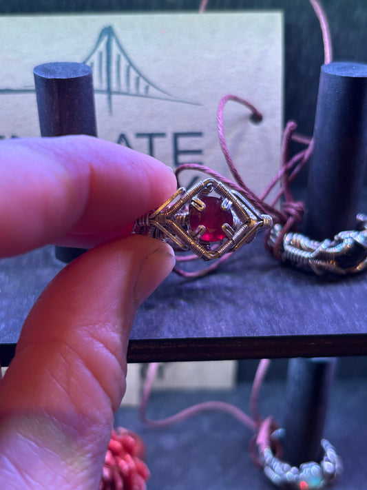 Hand holding a gold ring with a red gemstone against a blurred background