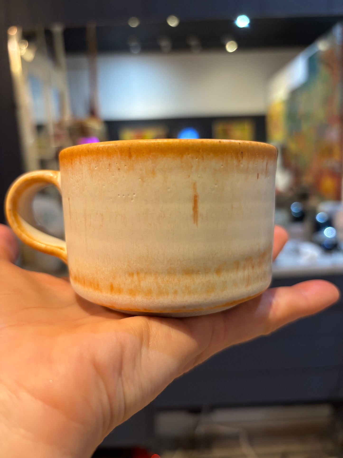 Hand holding a ceramic mug with orange and white design in a blurred indoor setting
